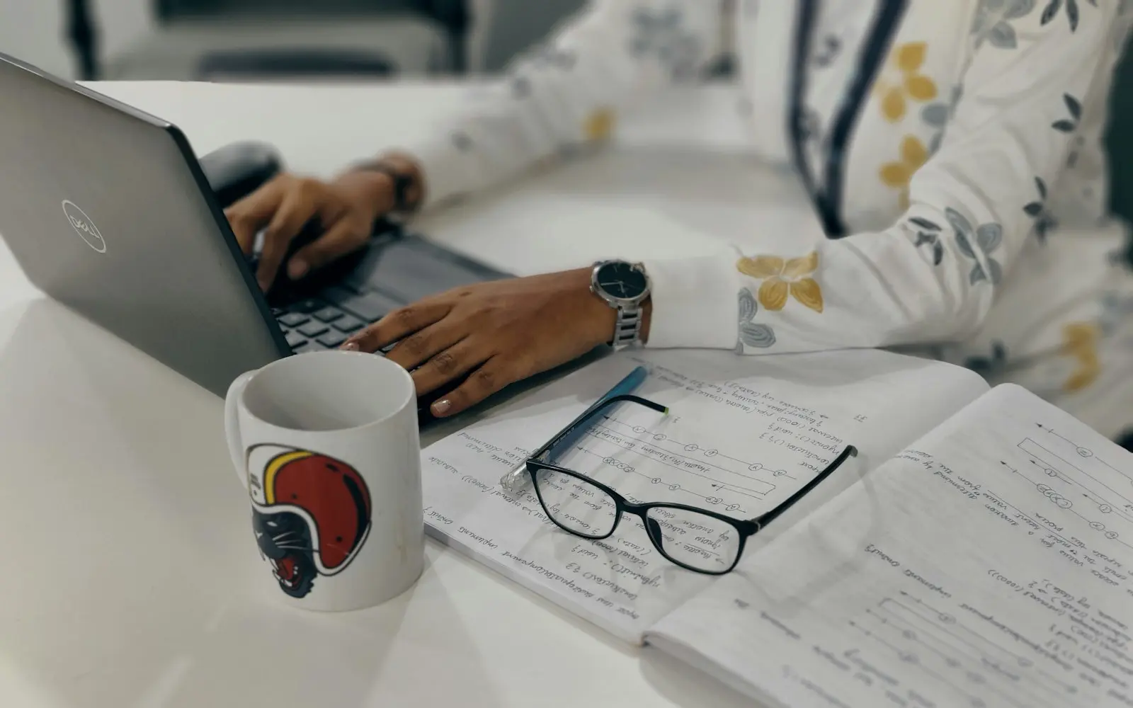 Writer typing on a laptop beside handwritten notes, glasses and a mug