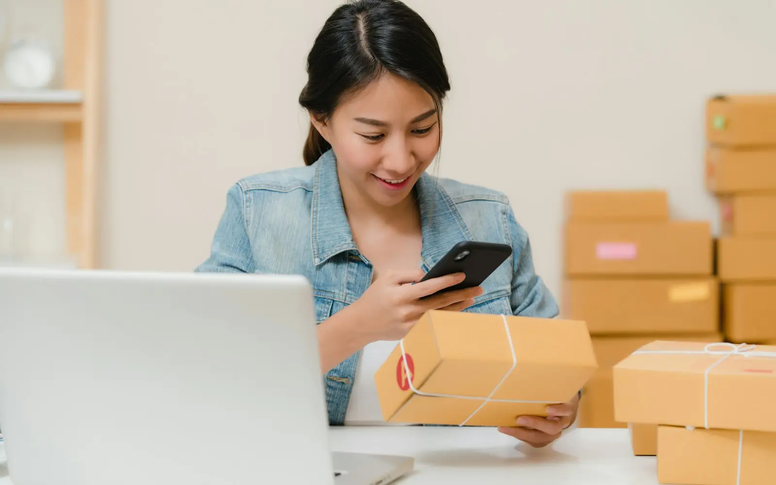Woman scanning a parcel with her phone beside a laptop and stacked boxes