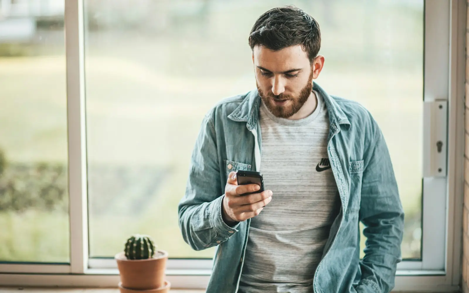 Man in a denim shirt reading his phone beside a bright window
