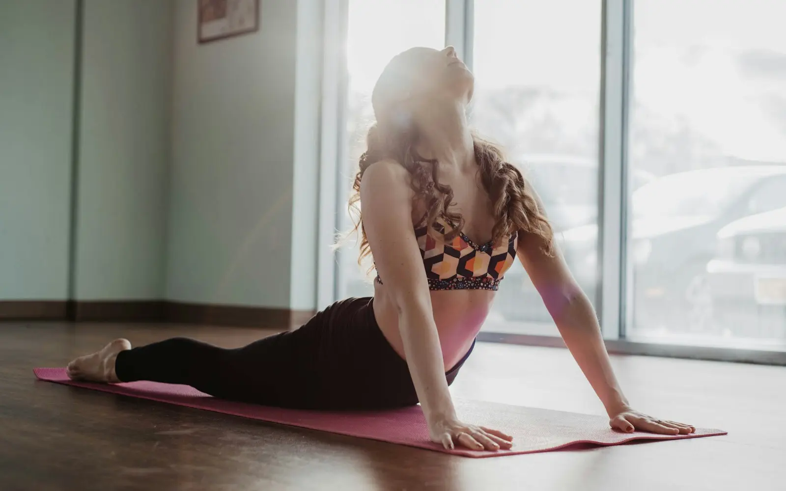 Woman stretching on a pink yoga mat in a bright studio beside large windows