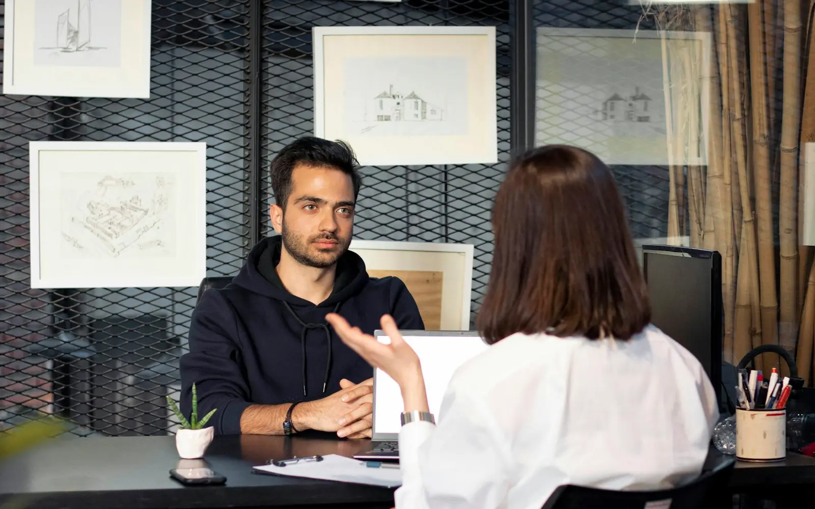 Man in a dark hoodie talking with a woman across a desk in a one-to-one meeting