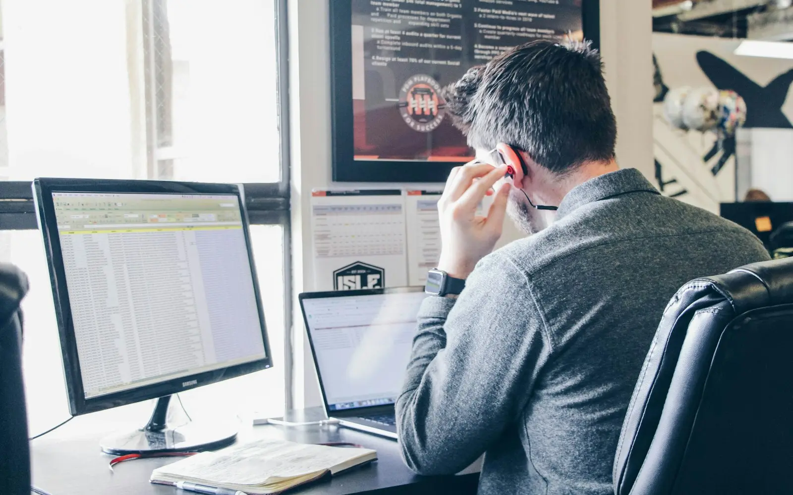 Man reviewing a spreadsheet on a desktop monitor beside a laptop in a busy office