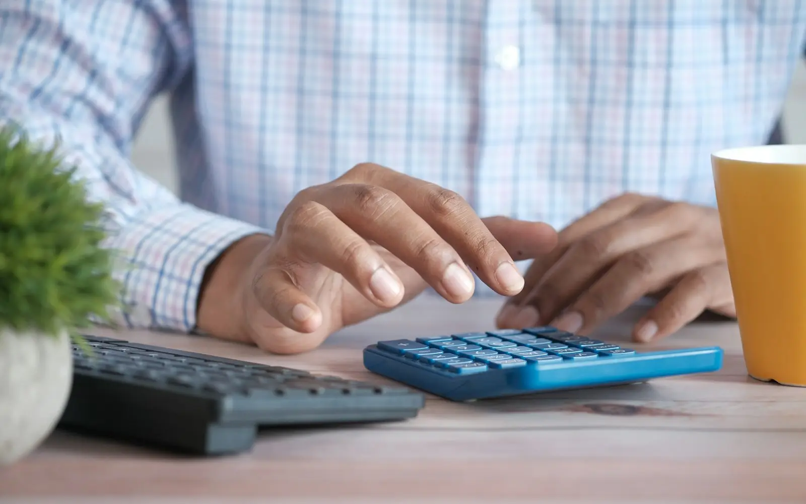 Hands using a calculator at a desk with a keyboard and coffee mug