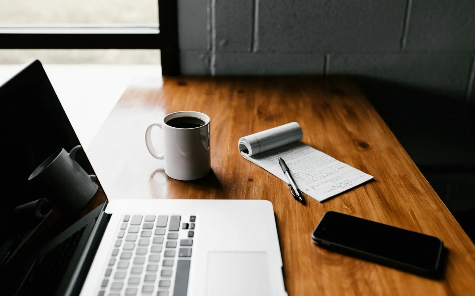 Laptop, notepad, pen and phone on a wooden desk beside a coffee mug