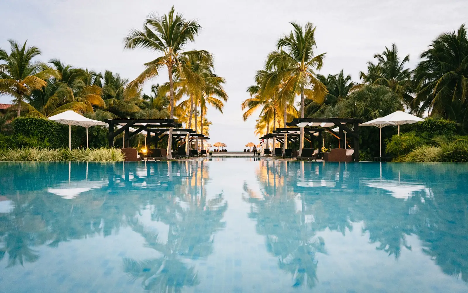 Resort swimming pool reflecting palm trees and shaded pavilions